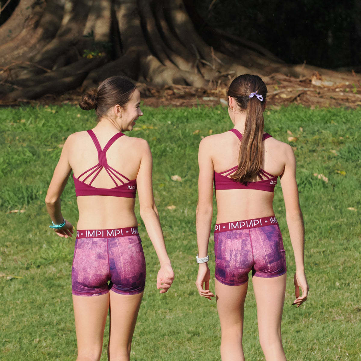 Two women in matching purple athletic wear walking on grass with a large tree in the background.